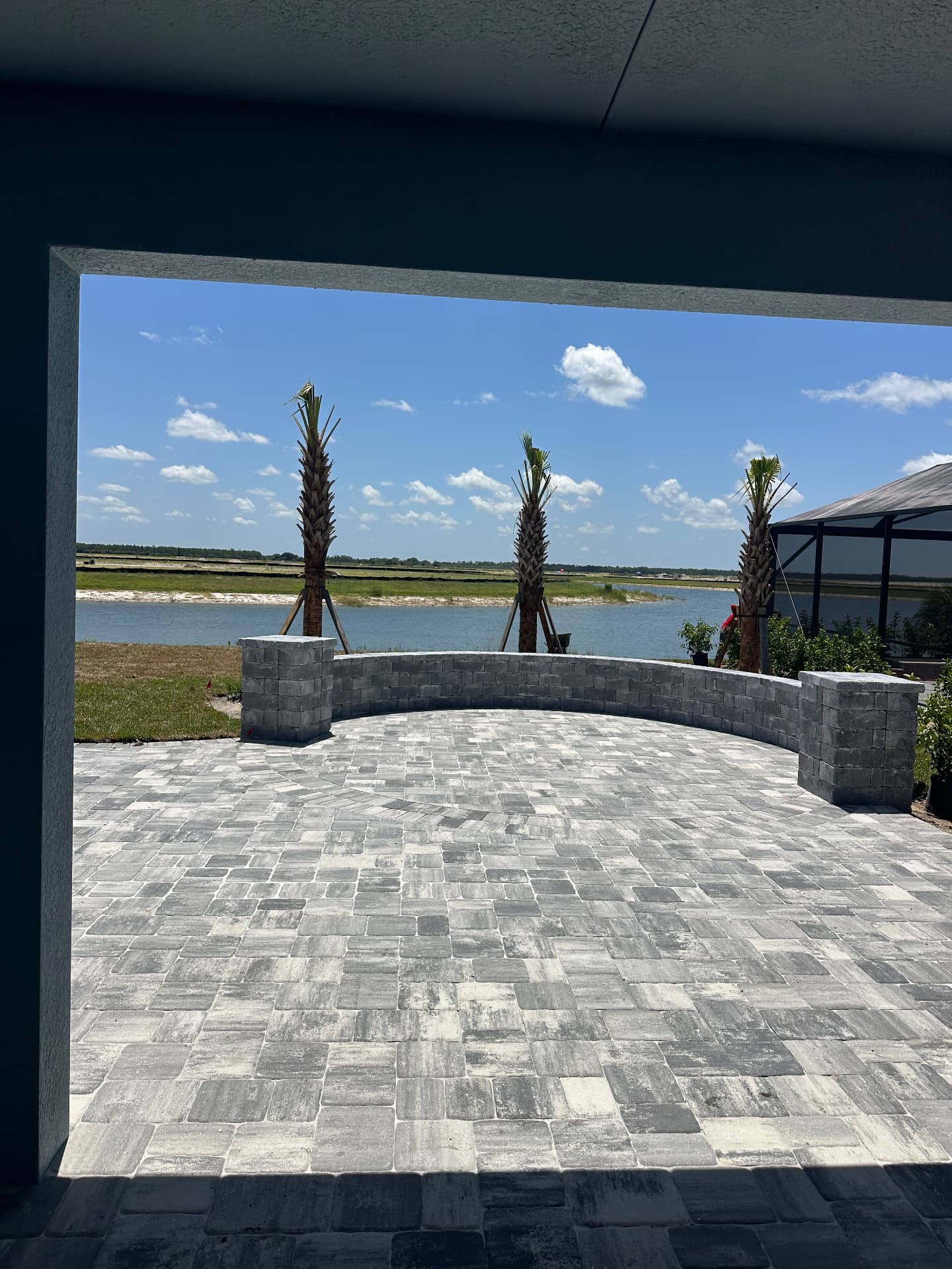Covered lanai looking out to a paver patio, seat wall, palms and lake in Kingston, Estero, Florida — August 2026