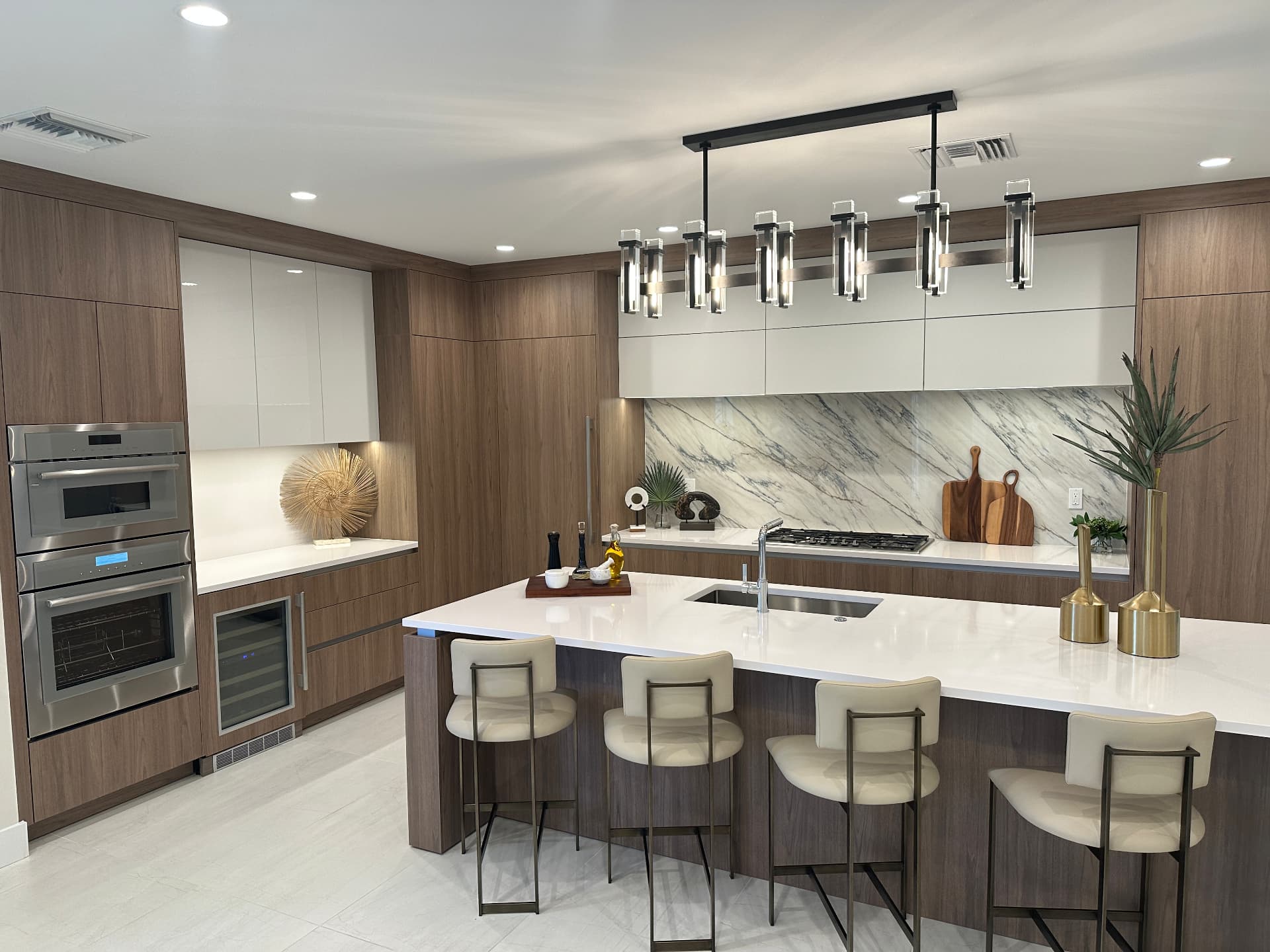 Model kitchen at The Island at West Bay Club in Estero, FL with walnut flat-panel cabinetry, marble slab backsplash, waterfall quartz island and built-in wall ovens, photographed August 2026